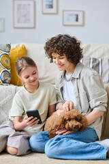 Vertical image of little girl showing photos on smartphone to her mom while they sitting with dog in living room