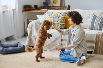 Young smiling woman training her dog in the room and giving her food for obedience