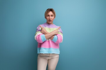 close-up of a charming blond young woman in a striped sweater in surprise showing her hands to the side on a blue background with copy space