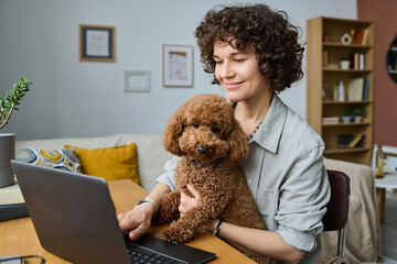 Young woman sitting at table and hugging her dog while working online on laptop in the room