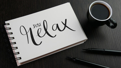 JUST RELAX black lettering in notebook with cup of coffee and pens on black wooden desk