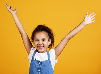 Studio, portrait and excited child with hands up and smile on face on yellow background. Young girl...