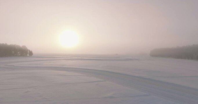 Aerial view following Lapland ice lake drifting track towards glowing misty sunrise horizon