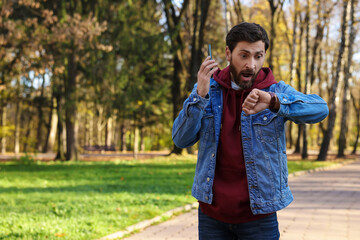 Emotional man talking by smartphone and checking time on watch in park, space for text. Being late concept