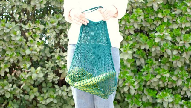 Female hand holding reusable shopping bag with vegetables on green background, zero waste concept.