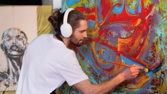 Creative innovative talented artist with long hair in headphones, wearing a black apron smeared with paint, creates masterpiece using red blue yellow paints, painting large canvas in creative studio.