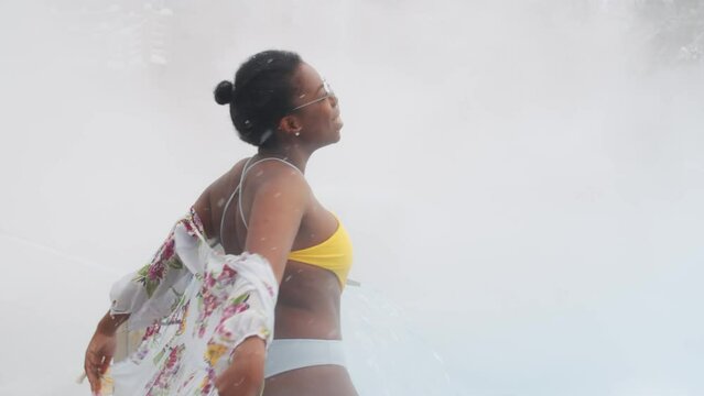 Medium portrait shot of charming African American woman wearing stylish swimsuit, cover-up and sunglasses standing against hot springs pool on snowy day posing on camera