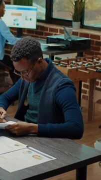 Vertical video: Corporate worker reviewing paperwork in office, looking at documents with statistics files in coworking space. African american man working on business development with data analytics.