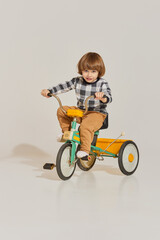 Charming cute boy with long hair riding retro bike and looking at camera over light background