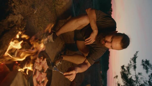 Young hipster man in shorts and gumshoes is cooking dinner on fire while sitting on log by the river and laughing. Vertical video