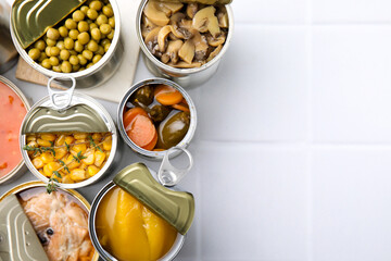 Open tin cans with different food on white tiled table, flat lay. Space for text