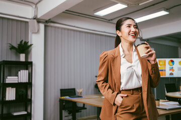 Portrait of a smart Asian young entrepreneur woman laughing and standing while holding a paper coffee cup in the office. Businesswoman wearing a blazer with a smile. Image with copy space.