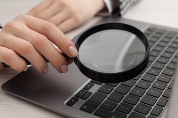 Woman holding magnifier near laptop at wooden table, closeup. Online searching concept