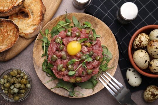 Tasty beef steak tartare served with yolk, capers and bread on brown table, flat lay