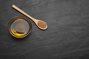 Bowl of natural oil and spoon with mustard seeds on black table, above view. Space for text