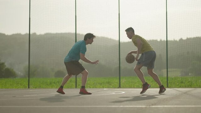 Slow motion - Two male basketball players playing basketball outside on a basketball court on a sunny day. A guy dribbles past his opponent and scores a layup in the red rim on the hoop to get a point