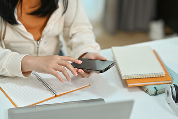 Young student woman sitting at desk and searching online information on smart phone. Cropped shot