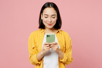 Young fun woman of Asian ethnicity wear yellow shirt white t-shirt hold in hand use mobile cell phone in green case isolated on plain pastel light pink background studio portrait. Lifestyle concept.