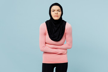 Young asian muslim fitness trainer sporty woman wear pink abaya hijab spend time in home gym suffer from stomach-ache griping bellyache isolated on plain blue background Workout sport fit abs concept