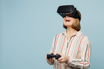Elderly happy woman 50s years old wear striped shirt hold in hand play pc game with joystick console watching in vr headset pc gadget isolated on plain pastel light blue cyan color background studio.