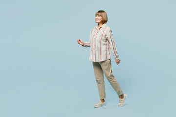 Full body side profile view smiling happy elderly woman 50s years old wear shirt walking going strolling isolated on plain pastel light blue cyan color background studio portrait. Lifestyle concept.