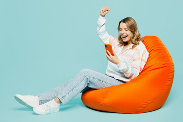 Full body smiling happy fun young woman wears striped hoody sit in bag chair hold in hand use...
