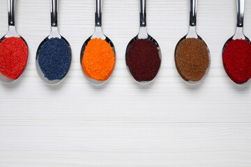 Spoons with different food coloring on white wooden table, flat lay. Space for text