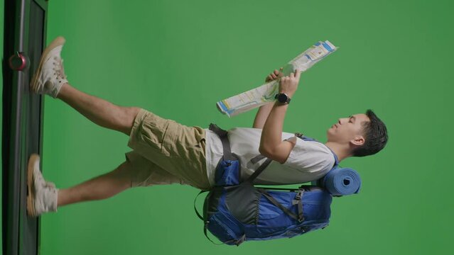 Full Body Side View Of Asian Male Hiker With Mountaineering Backpack Looking At The Map While Walking On A Treadmill On Green Screen Background In The Studio
