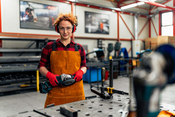 A young girl with curly red hair who is an apprentice in a metal workshop is using tools, she is...