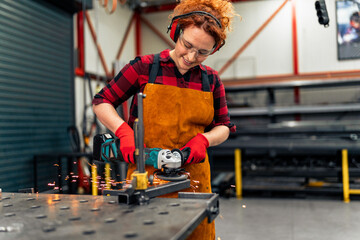 A young girl who is an apprentice in a metal workshop uses a grinder and works on a metal bar, she...