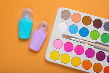 Watercolor palette with brush and markers on pale orange background, flat lay