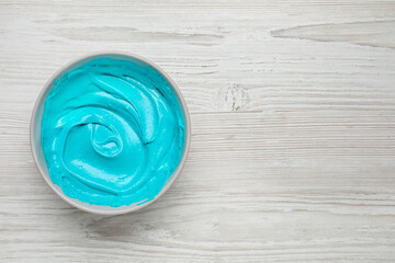 Bowl of cream with light blue food coloring on white wooden table, top view. Space for text