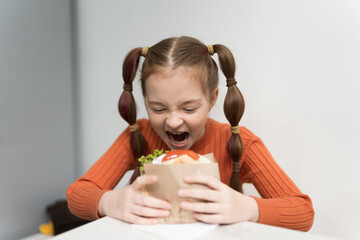 Funny little girl with ponytails biting a sandwich with a grin on her face. Cute hungry kid eating a pita souvlaki snack in a Greek restaurant
