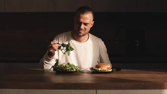 Dissatisfied male look with aversion on salad, he point on hamburger, choice and love it. Frowning man make choice salad or burger, unhealthy or healthy food, diet.