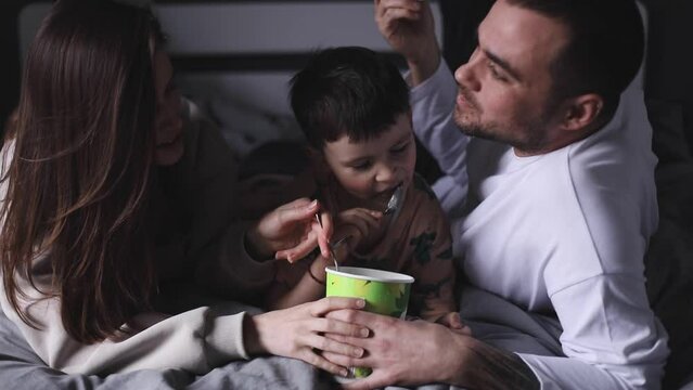 Cheerful family eating pistachio ice cream lying in the bed at home. Parents and kid eating by spoon ice cream. Family have fun at home. Love family concept at home. Parents kissing.