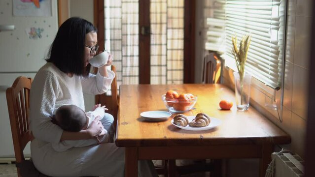 Classic home scene of a woman drinking coffee while breastfeeding her child in the kitchen. The best healthy nutrition for a baby