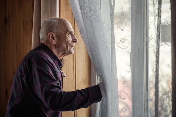 Single elderly man looking through window glass standing at home. Grandpa holding white curtain with hand and look out view from window. - obrazy, fototapety, plakaty