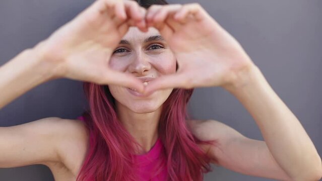 Happy attractive pink hair woman look camera send blow air kiss showing shape heart with hands, girl wear pink top isolated on grey background. Love, romantic concept.