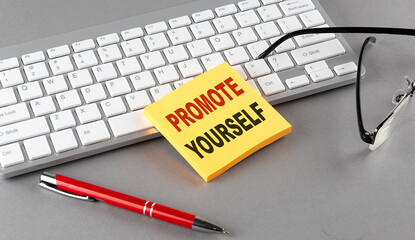PROMOTE YOURSELF text on a sticky with keyboard, pen glasses on grey background