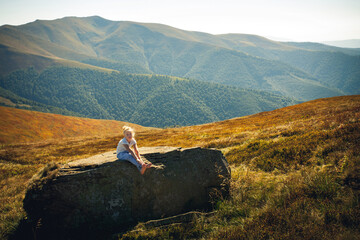 A little girl in blue pants and a light T-shirt sits on a big stone in the mountains, hot summer. Around the grass, trees and blue sky with clouds.