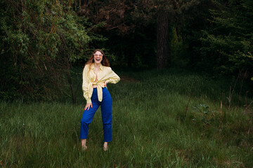 A smilling girl with blond curly hair dressed in blue jeans and a yellow shirt, wearing sunglasses, stands in a picturesque park in summer.