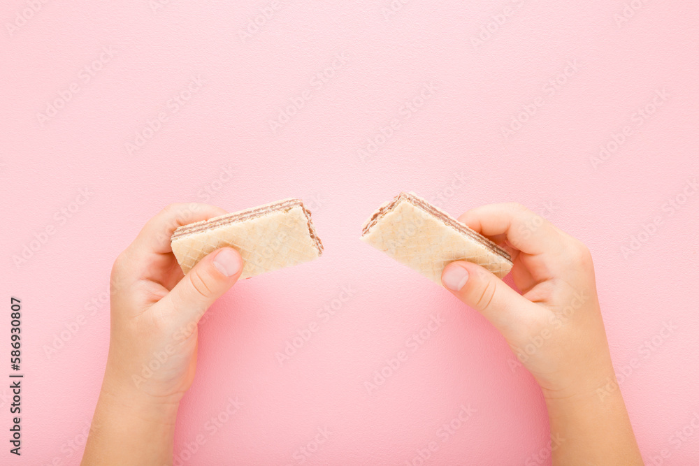 Wall mural little girl hands holding broken wafer pieces on light pink table background. pastel color. sweet sn