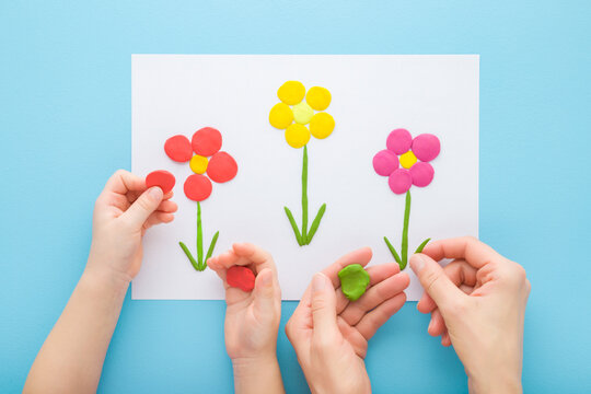 Adult mother and child hands modeling colorful flowers from clay on white paper on blue table background. Pastel color. Playing and spending time together. Point of view shot. Closeup. Top down view.