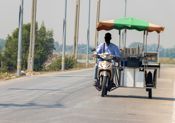 An ice cream vendor rides a three-wheeled motorcycle along a village road, Thailand
