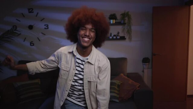 Charismatic and joyful afro-american young man dancing in the living room, having fun alone at home. Happy looking at camera.