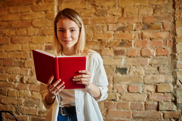 Positive woman reading book near brick wall