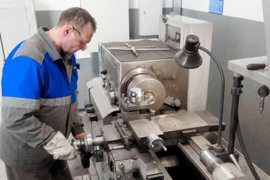 Elderly turner of 50-55 years old in overalls works at lathe in production shop. Processing of metal products..