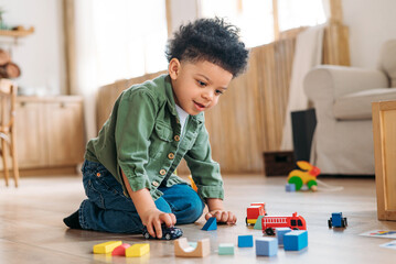 Concentrated little african american boy playing toys sitting on warm floor in modern living room. Baby development. Small tower. Learning creative concept