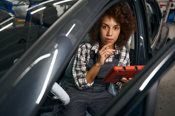 Woman auto mechanic sits in car interior with a device