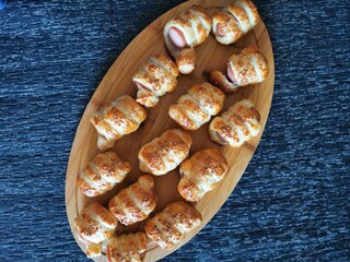 Baked and sesame-sprinkled sausages in dough, lying on a wooden oval board.